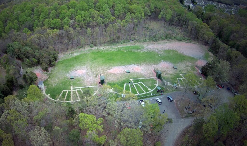 Drone photo looking down on the fan-shaped skeet and trap shooting stations, with the shot-fall field and tree line beyond